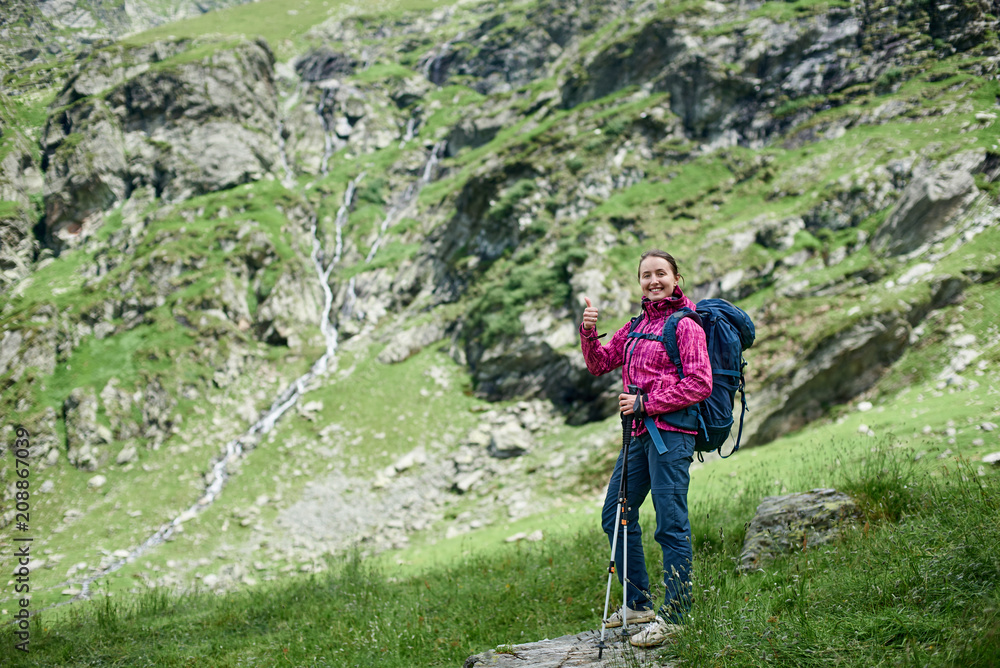 Young happy famele hiker with trekking sticks and a backpack is standing on a stone at the foot of the Fagaras Mountains, Romania. Girl smiling to the camera and showing thumbs up Young happy famele hiker with trekking sticks and a backpack is standing on a stone at the foot of the Fagaras Mountains, Romania. Girl smiling to the camera and showing thumbs up