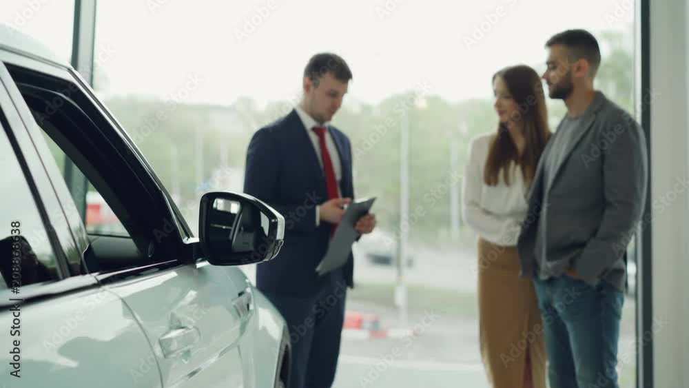 Professional automobile seller in suit is having conversation with attractive rich couple telling them about new car model, cheerful buyers are smiling. Focus on beautiful auto. Professional automobile seller in suit is having conversation with attractive rich couple telling them about new car model, cheerful buyers are smiling. Focus on beautiful auto.