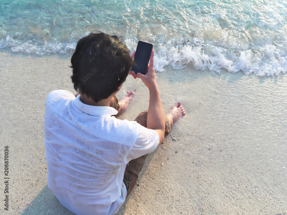 Young Asian man using mobile smart phone on the sandy beach. Summer vacations concept.