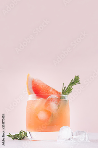 Fresh grapefruit juice with ice, piece and green rosemary closeup on soft white wood table and pink background, vertical.