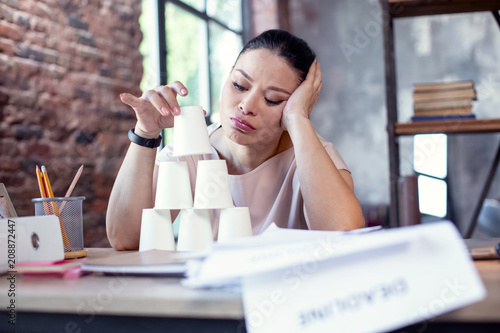 Play at work. Low angle of dreary female freelancer using paper cups and sitting at table photo