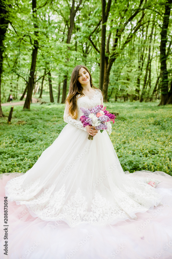 A sweet bride in a white dress is in the park with a bouquet in her hands