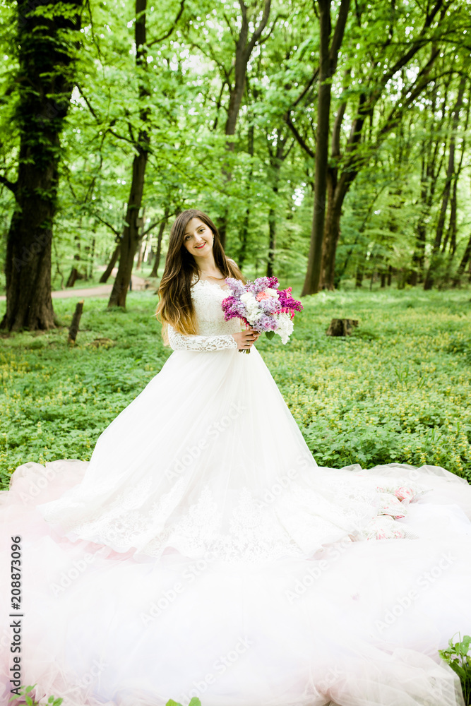 A pretty girl in a white dress is in the park with a bouquet in her hands