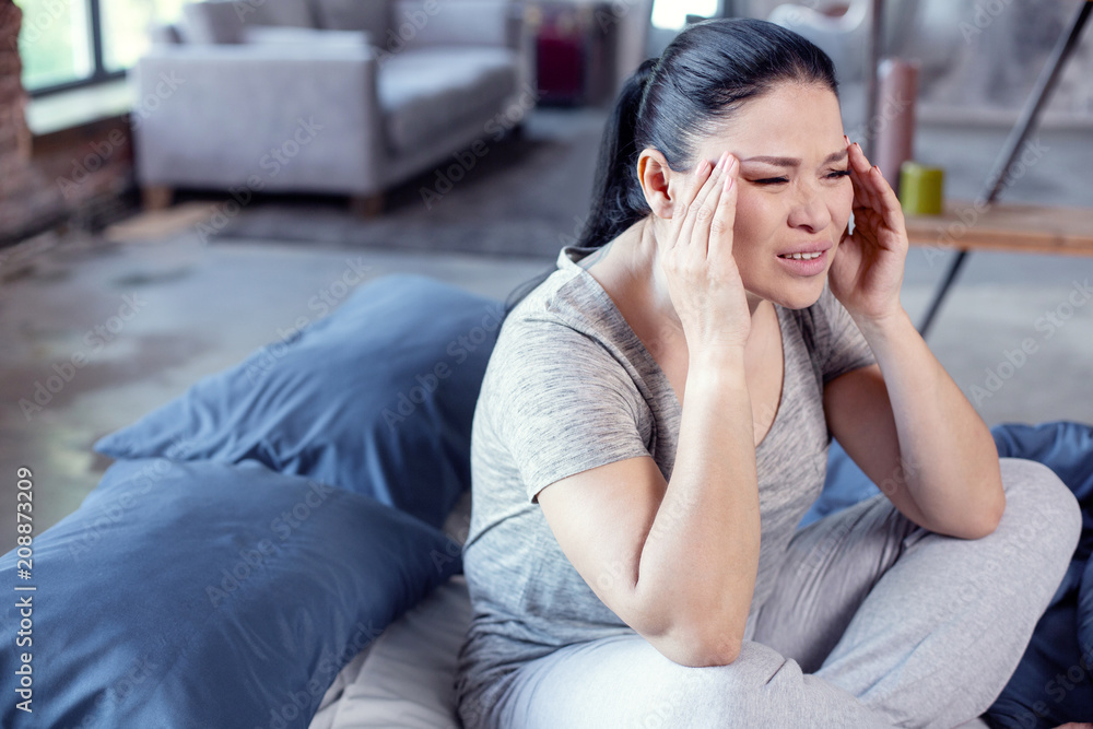 Obraz premium Symptom of disease. Tormented worn woman sitting on bed and touching temples