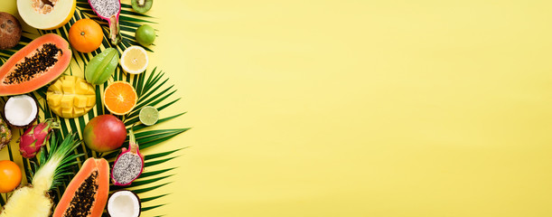 Exotic fruits and tropical palm leaves on pastel yellow background - papaya, mango, pineapple, banana, carambola, dragon fruit, kiwi, lemon, orange, melon, coconut, lime. Banner. Top view.