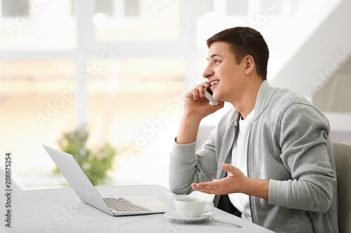 Young man talking on phone while working with laptop in cafe