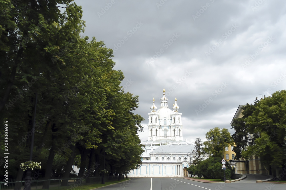 Fototapeta premium road past the park to the elegant white church, Smolny Convent site, Saint Peterburg (Russia); stormy clouds in the sky