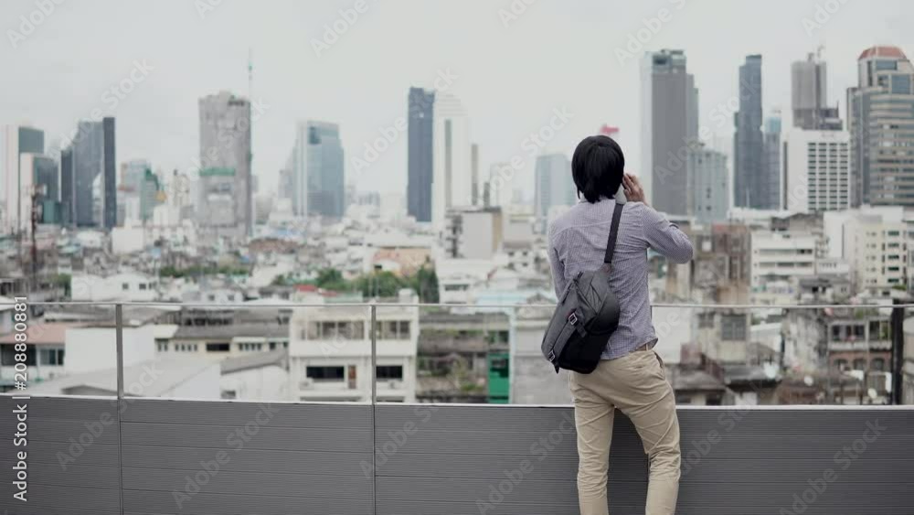 Young Asian businessman using smartphone for business talk standing on office building rooftop terrace with city view in the background. Business communication and corporation concepts. 4K video Young Asian businessman using smartphone for business talk standing on office building rooftop terrace with city view in the background. Business communication and corporation concepts. 4K video