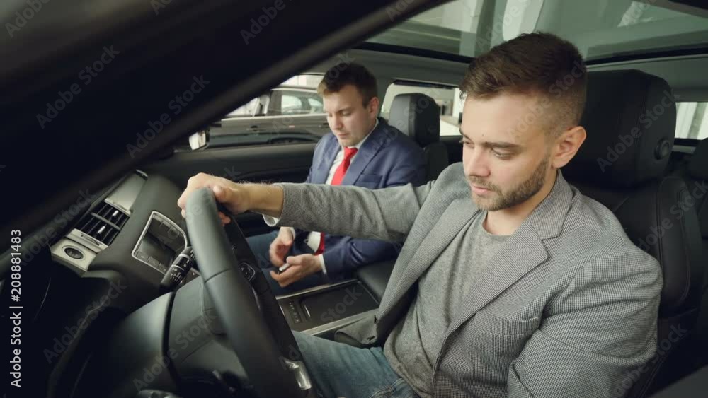 Bearded guy is discussing car features with salesman sitting together inside beautiful expensive car and talking. Buyer is holding steering wheel and touching car parts. Bearded guy is discussing car features with salesman sitting together inside beautiful expensive car and talking. Buyer is holding steering wheel and touching car parts.