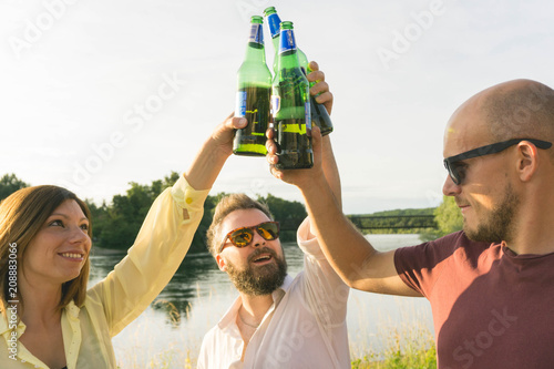 Group of happy friends drinking and toasting beer bottles at river at sunset. Friendship concept with young people having fun together at nature. Group of friends having drinks together.