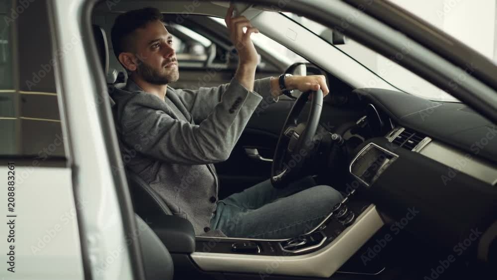 Serious young man is sitting inside new car in motor showroom and checking electronics pressing buttons on control panel touching car parts. Purchasing automobile concept. Serious young man is sitting inside new car in motor showroom and checking electronics pressing buttons on control panel touching car parts. Purchasing automobile concept.