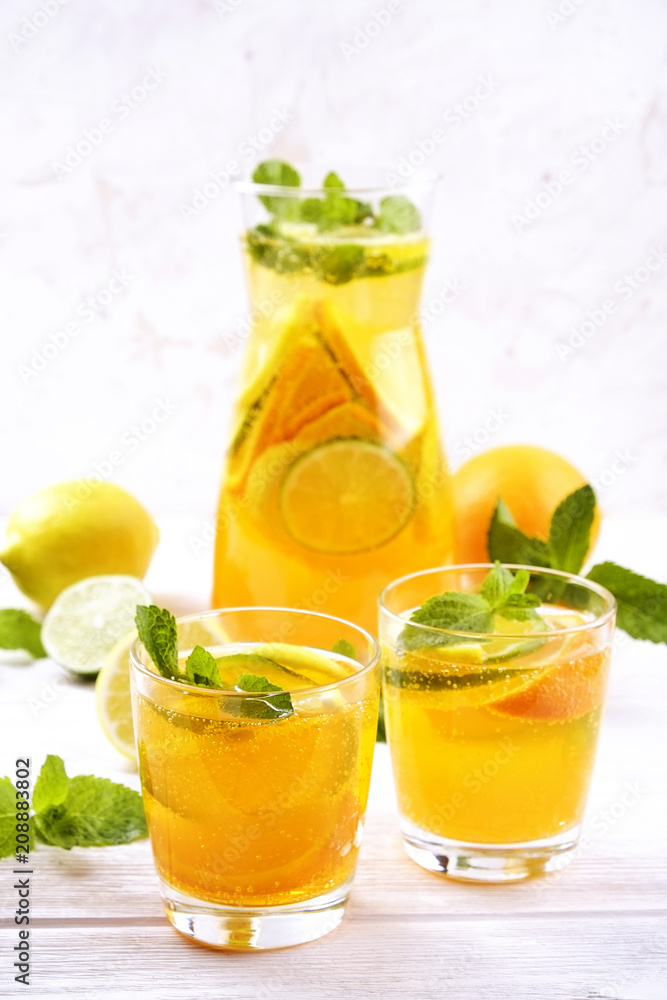 Two glasses & pitcher bottle of homemade refreshing lemonade with slices of organic ripe lemon, orange and lime, mint & ice on rustic white wooden table background. Close up, top view, copy space. Two glasses & pitcher bottle of homemade refreshing lemonade with slices of organic ripe lemon, orange and lime, mint & ice on rustic white wooden table background. Close up, top view, copy space.