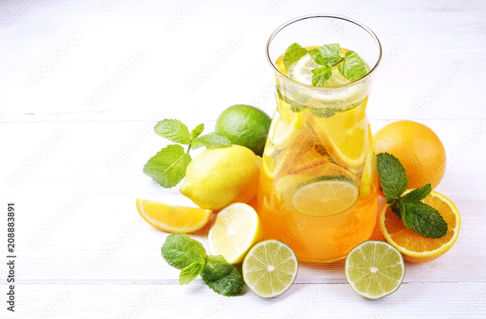 Fototapeta premium Two glasses & pitcher bottle of homemade refreshing lemonade with slices of organic ripe lemon, orange and lime, mint & ice on rustic white wooden table background. Close up, top view, copy space.
