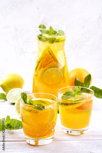 Two glasses & pitcher bottle of homemade refreshing lemonade with slices of organic ripe lemon, orange and lime, mint & ice on rustic white wooden table background. Close up, top view, copy space.