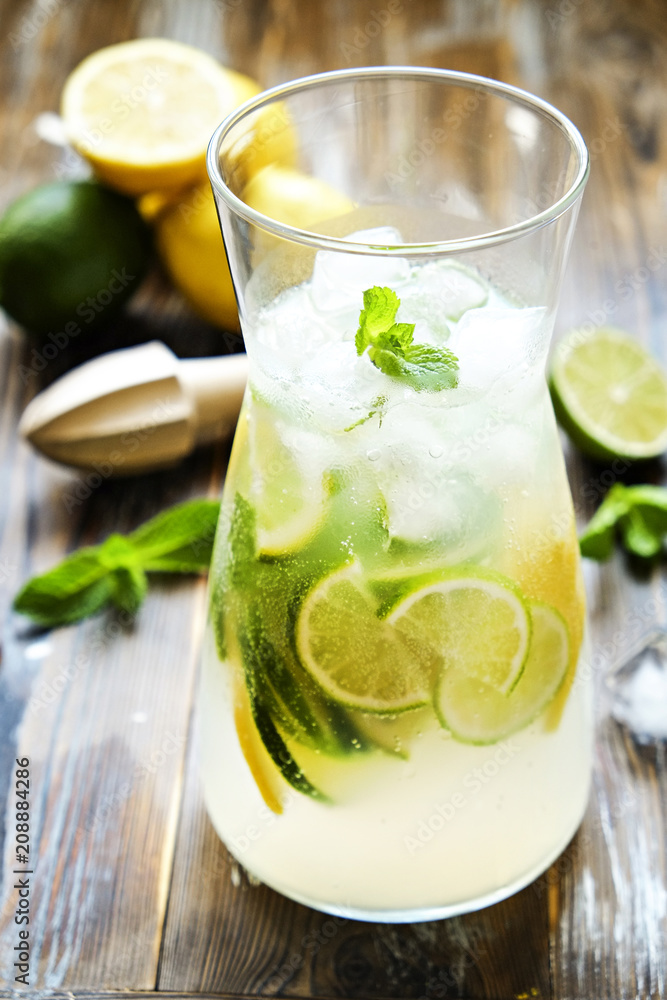 Iced lemonade pitcher, wooden juicer, cold citrus infused water beverage with lemon & lime slices, mint leaves on brown grunged wood textured table sunny day, grunged background. Close up, copy space. Iced lemonade pitcher, wooden juicer, cold citrus infused water beverage with lemon & lime slices, mint leaves on brown grunged wood textured table sunny day, grunged background. Close up, copy space.