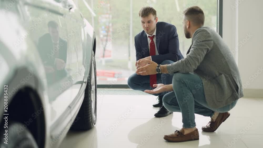 Two men cheerful customer and friendly salesman are talking and gesturing discussing car model while squatting near expensive automobile. Buying and selling vehicles concept. Two men cheerful customer and friendly salesman are talking and gesturing discussing car model while squatting near expensive automobile. Buying and selling vehicles concept.