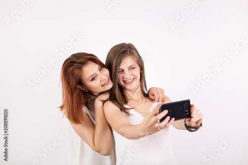 Portrait of two gorgeous young woman, a redhair and a brown-haired, taking a selfie over a white wall in studio