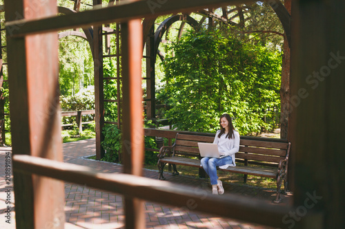 Young successful businesswoman in light casual clothes. Woman sitting on bench working on modern laptop pc computer in city park in street outdoors on nature. Mobile Office. Freelance business concept