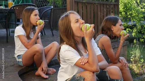 Eating biting green color ripe apples fruits triple sisters twins teen girls sitting on backyard porch terrace