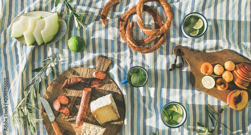 Flat-lay of summer picnic set with fruit, cheese, sausage, bagels and lemonade over striped blanket, top view