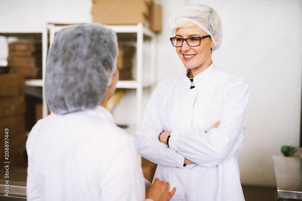 Two young joyful female workers in sterile cloths are having a chat while standing in factory cargo room.