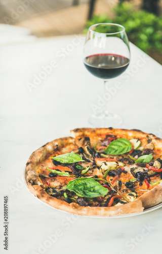 Summer dinner or lunch. Freshly baked Italian vegetarian pizza with vegetables, fresh basil and glass of red wine over white marble table, copy space, vertical composition