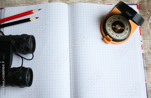 A top view of a set of objects for planning a trip, a tourist route or a hike. An open notepad with clean sheets, a compass, binoculars and two pencils of red and black colors. Copy space. Flat lay.