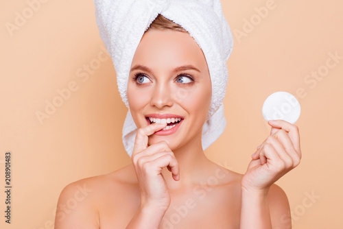 Portrait of dreamy minded ponder girl with turban on head holding using white cotton pad for removing makeup isolated on beige background