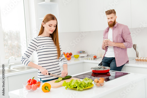 Psychology stress offense misunderstanding conflict crisis concept. Portrait of disappointed man drinking coffee while woman cutting vegetables for salad in modern white kitchen with interior