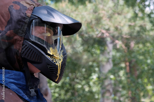 Yellow blot paint on a protective glass mask for playing paintball against the backdrop of the forest. Profile of a man in a black helmet with a visor and protective clothing.