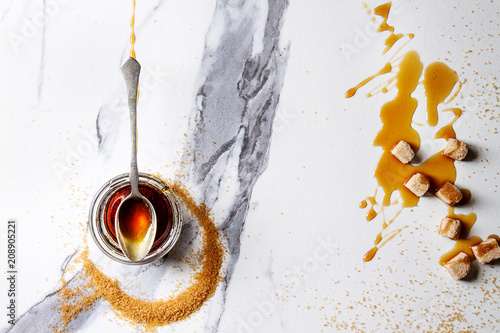 Konstfotografi Homemade liquid transparent brown sugar caramel in glass jar with spoon and cane sugar cubes over white marble background