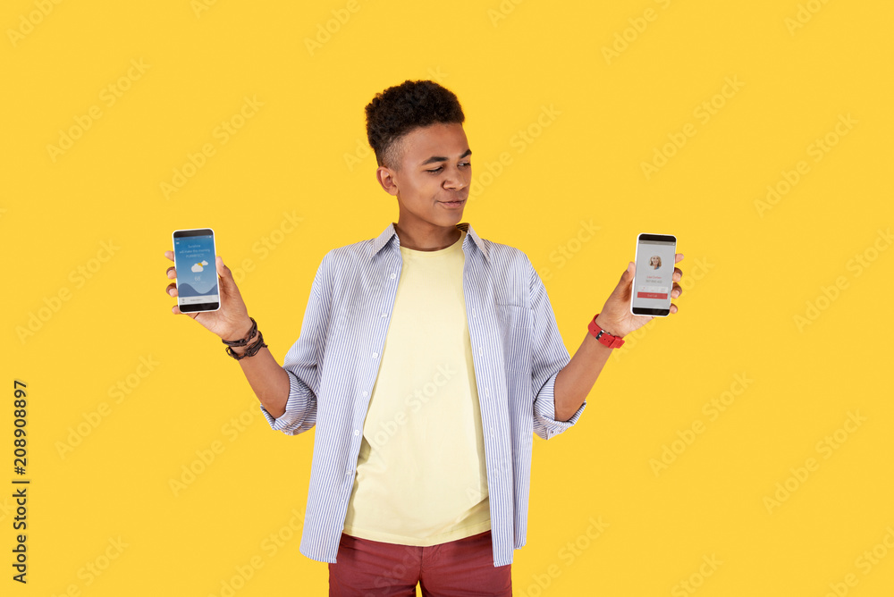 This is better. Smart young man standing against yellow background while making a choice between two smartphones This is better. Smart young man standing against yellow background while making a choice between two smartphones