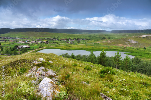 Heavy rain over the Ural village. Lake in the hills. Beautiful colorful sky. Russia