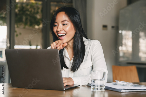 Wallpaper Mural Photo of cheerful asian woman 20s wearing white shirt laughing and pointing finger at screen of laptop, while speaking or chatting on video call in office Torontodigital.ca