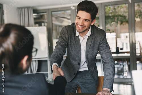 Business, career and placement concept - joyful young man 30s smiling and shaking hands with businesswoman, when was recruited during interview in office