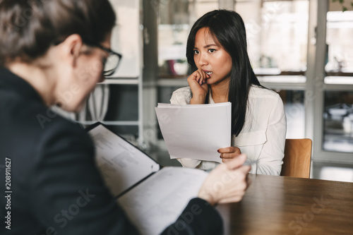 Image of concentrated tense asian woman looking at businesswoman, while sitting at table in office during job interview - business, career and recruitment concept photo