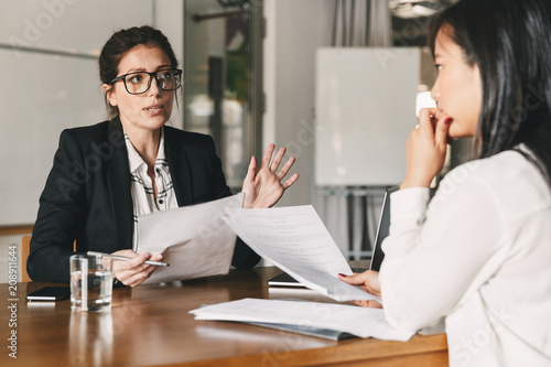 Photo of strict caucasian woman holding resume, and negotiating with female candidate during corporate meeting or job interview - business, career and placement concept