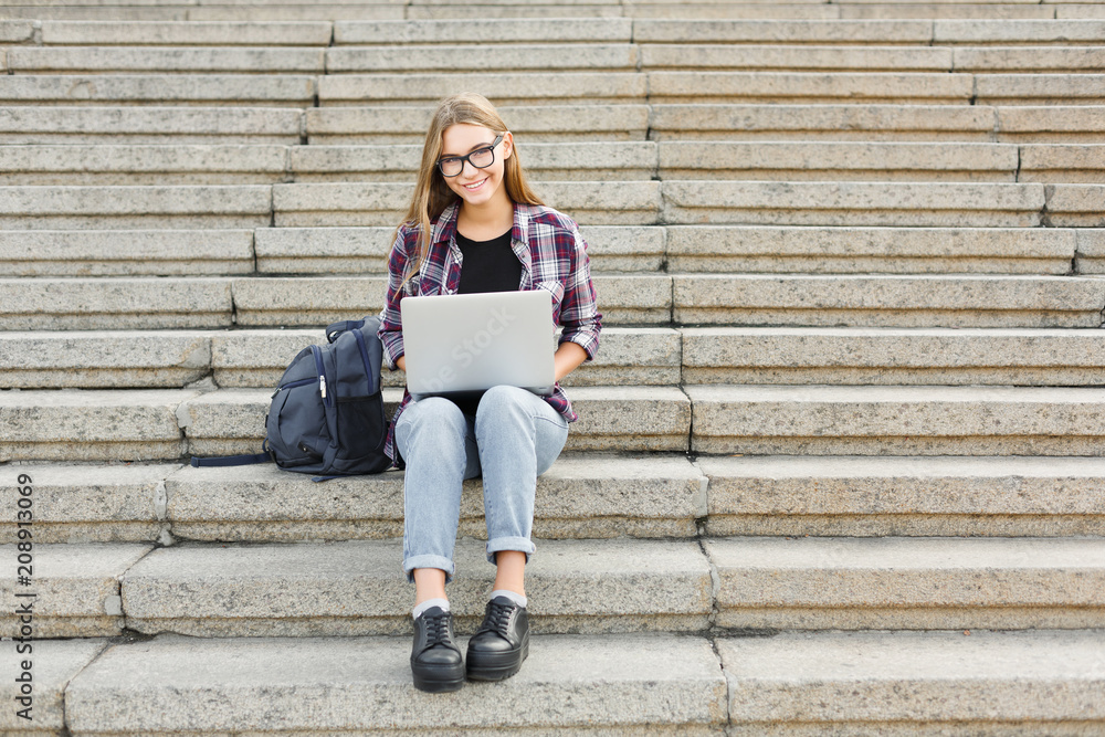 © Prostock-studio - Smiling student sitting on stairs using laptop