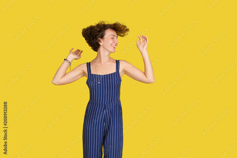 Joyful caucasian female model shaking head with brunette curly hair and smiles broadly, wearing trendy blue overall. Girl feels free and young, having fun against yellow studio wall background