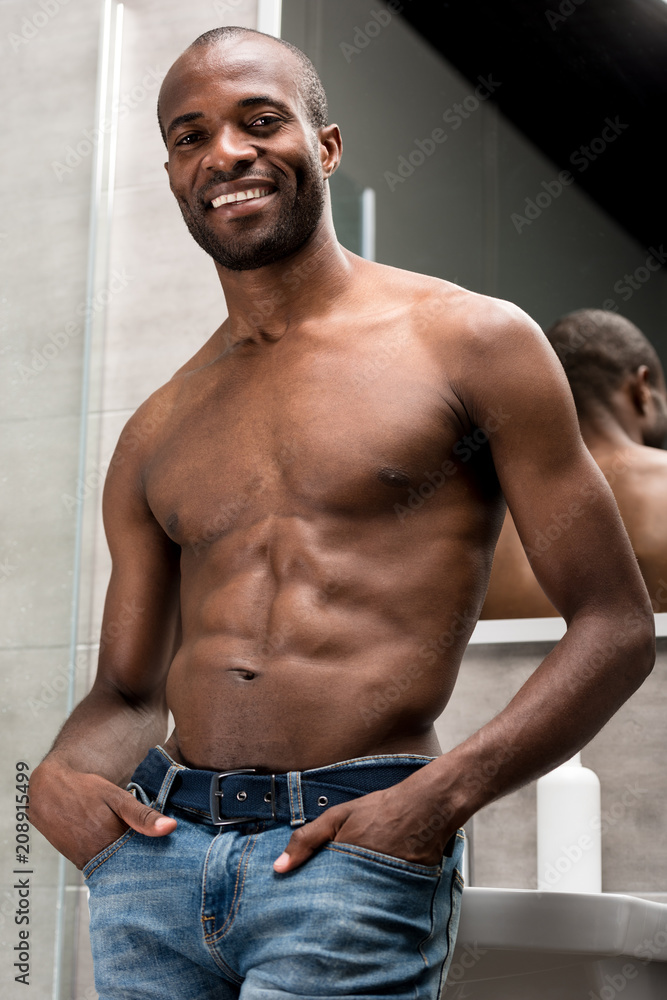 low angle view of handsome shirtless african american man standing with hands in pockets and smiling at camera in bathroom