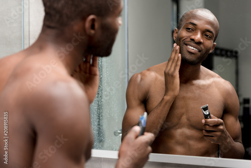 smiling african american man shaving with electric trimmer in bathroom photo