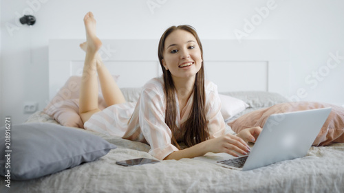 cheerful brown haired woman is lying on a bed in room in day with notebook in front of her and looking to a camera