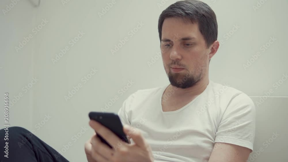 Grown man with beard lying in bed wearing white tshirt with a cellphone. Man using smartphone for social network indoor. Relaxing at home. Grown man with beard lying in bed wearing white tshirt with a cellphone. Man using smartphone for social network indoor. Relaxing at home.