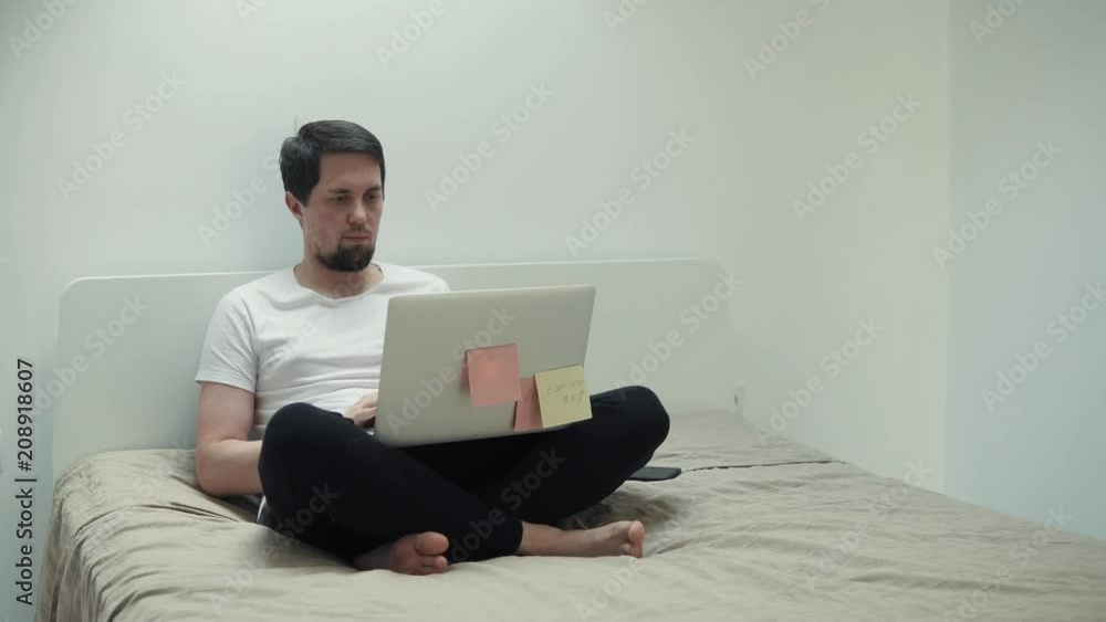 Relaxed grown man is lying in bed with a laptop covered in notes. Casual guy chatting with friends and typing messages online. Day indoor. Relaxed grown man is lying in bed with a laptop covered in notes. Casual guy chatting with friends and typing messages online. Day indoor.