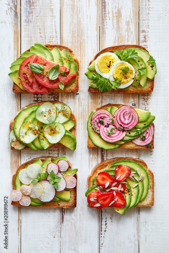 Avocado sandwiches, toasts with various vegetarian toppings on a white wooden table, top view