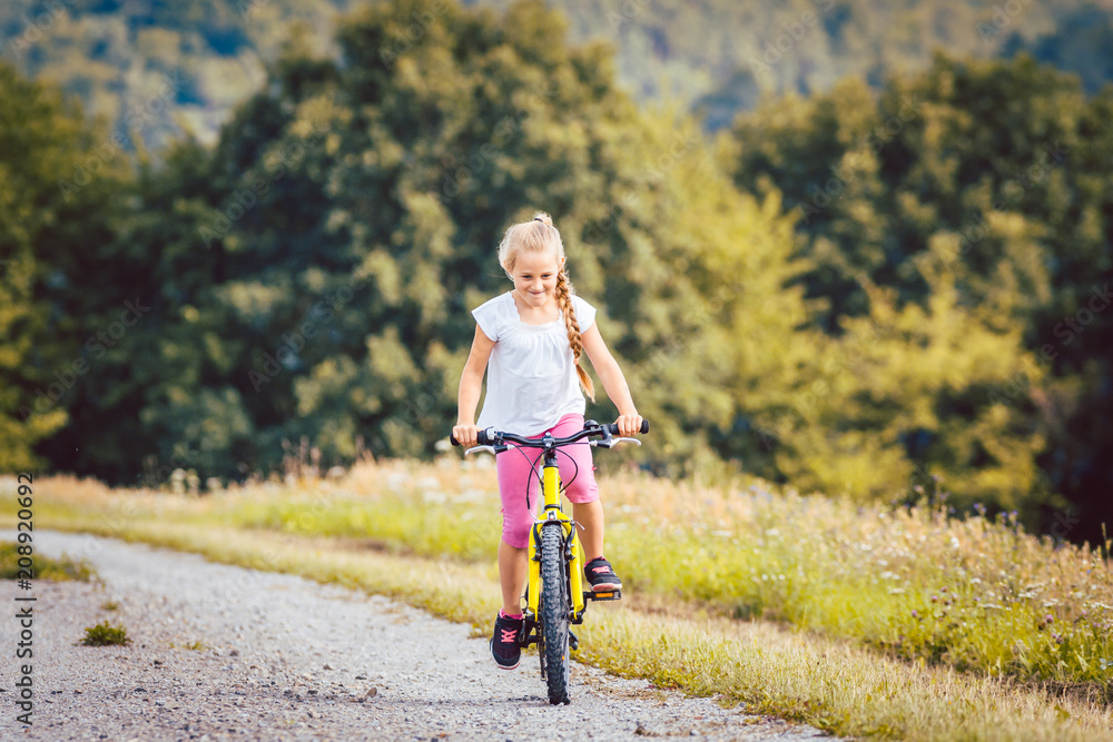 Fototapeta premium Girl on bicycle cycling down a dirt path in summer, woods in the background
