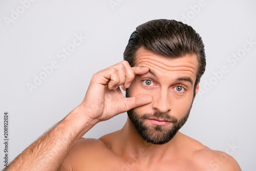 Muscular, manly, tired man open his eye widely with fingers, having redness inside because of sleep deprivation, showing his problem to oculist, isolated over grey background