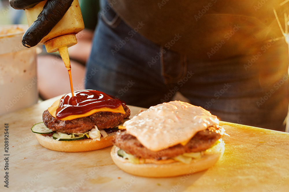 The chef prepares a burger in the open air kitchen. Dripping sauce. Street fast food. Sunset light.