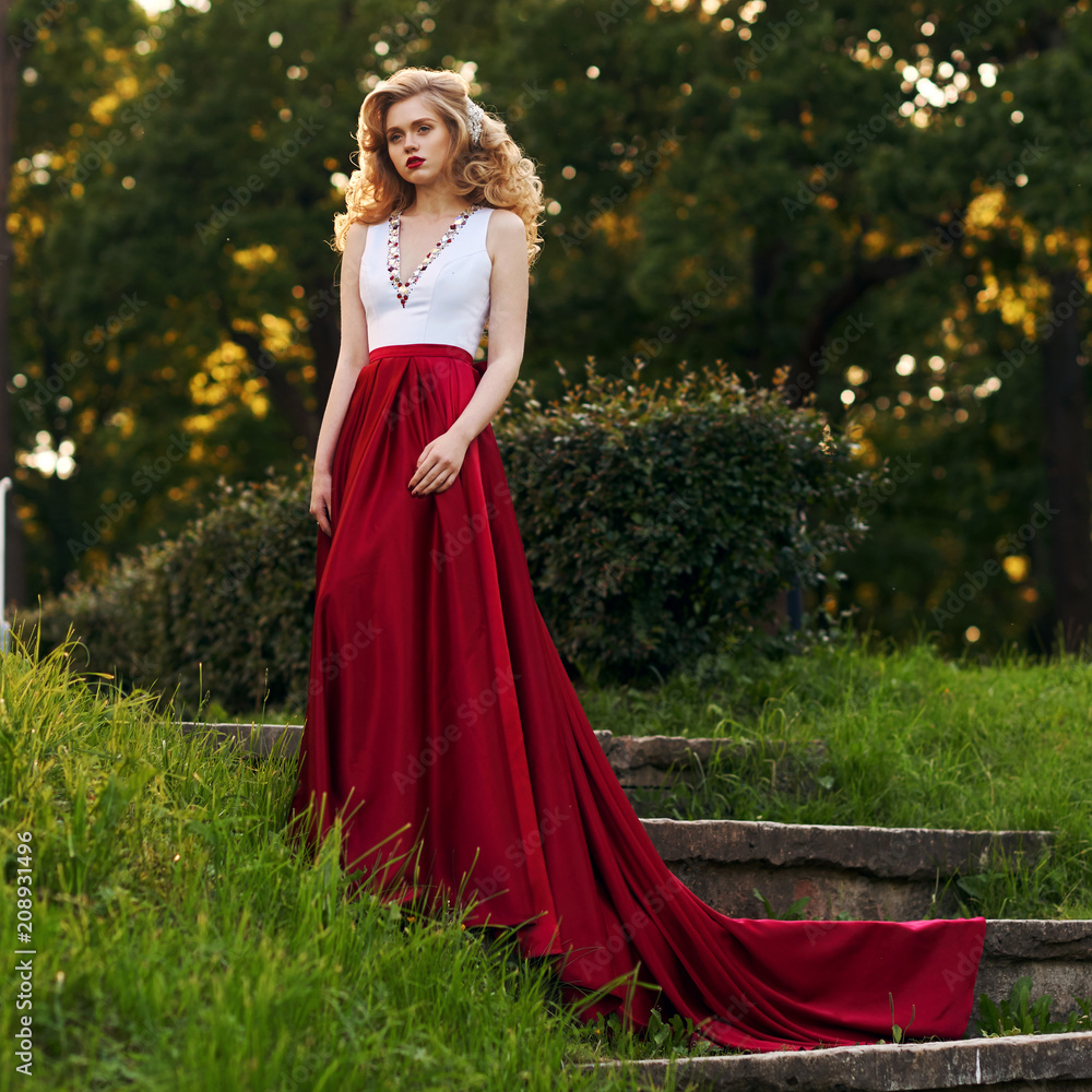 Fashion style photo of young beautiful woman in evening dress with white top and red flying skirt posing in summer park at sunset under green trees and close to old brick tower.