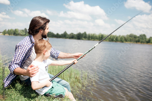A picture of boys sitting together at the edge of lake and fishing. Boy is holding long fish-rod while his dad is guiding him to fishing right.